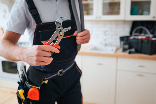 Cut view of man in working uniform hold wrench. He stand in kitchen. Daylight. Tools on belt. Toolbox on desk.