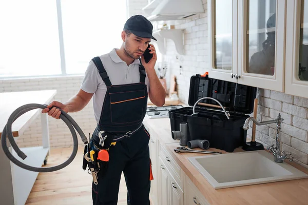 In der Küche telefonieren fleißige Handwerker. Er hält Schlauch. Werkzeugkiste auf dem Schreibtisch. Mann steht am Waschbecken. — Stockfoto