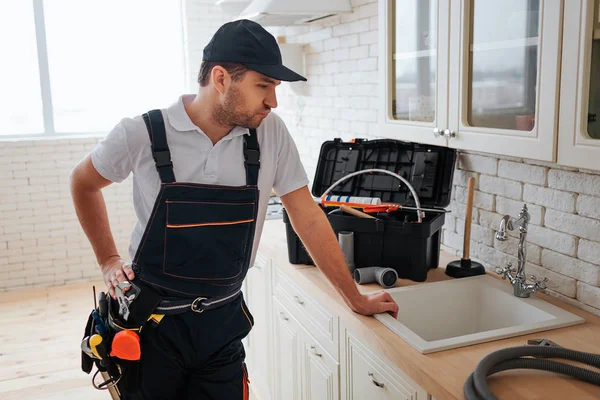 Nachdenkliche Handwerker stehen in der Küche am Spülbecken und betrachten es. Werkzeugkiste und Schlauch auf dem Schreibtisch. Mann trägt Uniform und Gürtel mit Werkzeug. — Stockfoto