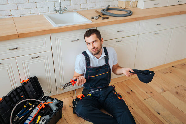 Handyman sit on floor in kitchen. He look on camera confusingly. Guy hold cap and wrench. Having rest.