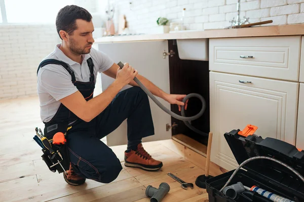 Konzentrierter junger Arbeiter, der in der Küche Reparaturen erledigt. Er sitzt in Kaderpose und hält Schlauch. Stück Rohr auf dem Boden. Werkzeugkiste geöffnet. — Stockfoto