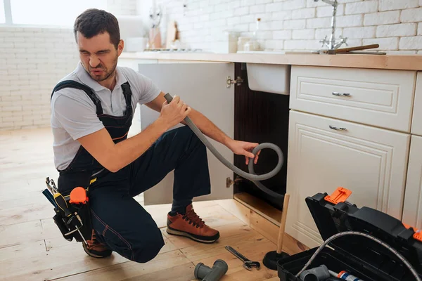 Junger Mann hält Schlauch in der Hand. Schlechter Geruch. Stinkt. Heimwerker leiden darunter. geöffneter Werkzeugkasten und ein Stück Rohr auf dem Boden. — Stockfoto