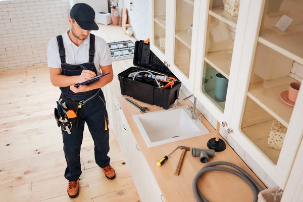 Klempner in der Küche. in Dokument schreiben. Typen stehen am Waschbecken. Werkzeuge und Instrumente auf dem ganzen Schreibtisch. Schlauch, Hammer, Roulette, Brustwarzen. — Stockfoto