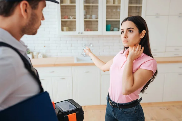 Femme concentrée sérieuse se tenir avec plombier dans la cuisine. Elle pointe du doigt l'évier. Le gars tient la boîte à outils et regarde dans cette direction . — Photo