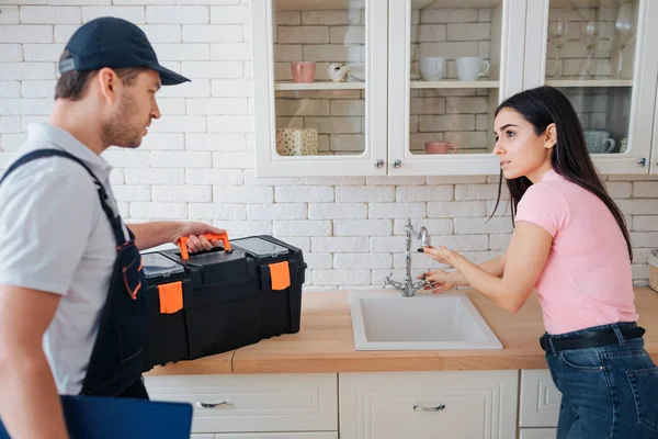 Young woman need help with sink in kitchen. She point on tap. Plumber stand beside her. He hold toolbox.