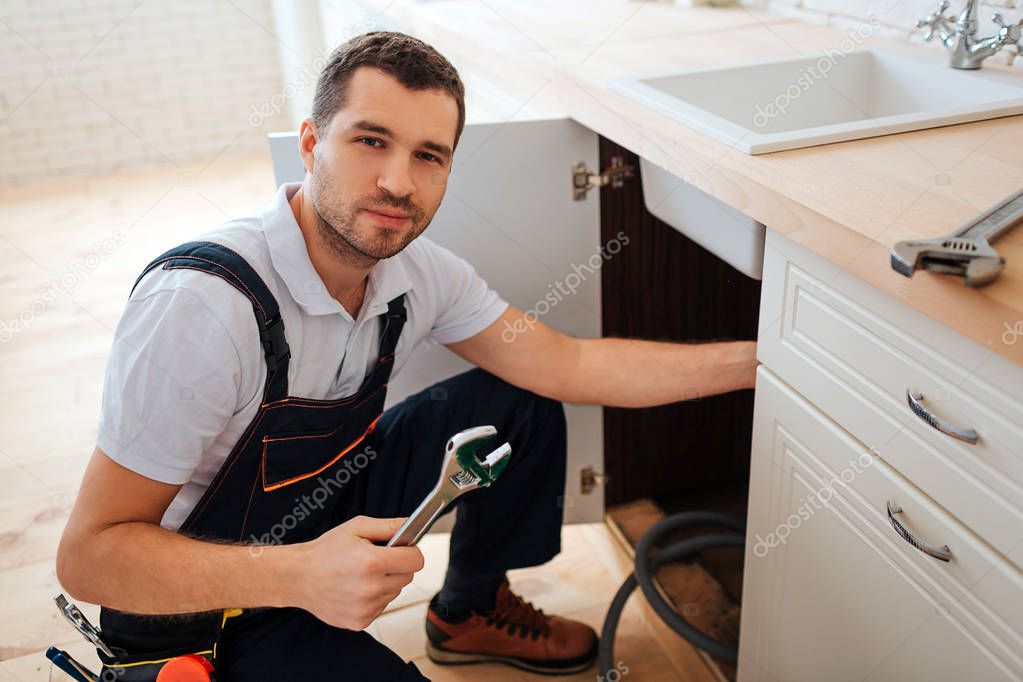 Hansome young plumber look on camera. He stand in kitchen and hold wrench. Repair. Another wrench lying on desk.