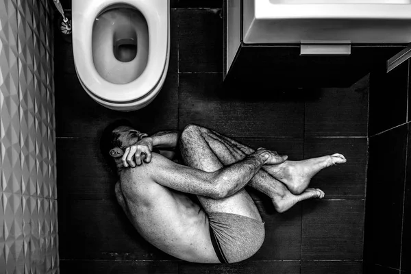 Ung man lida inne i övriga rum ensam. Han liggande på golvet och hålla benen med händerna. Guy slitage endast shorts. Öppnade upp i toilette. SmÃ¤rta och missbruk. Svart och vit bild. — Stockfoto