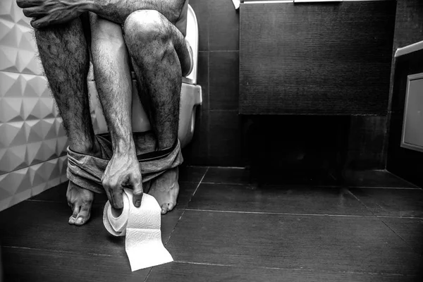 Cut view mans legs. Guy sit on toilet in rest room. He naked and have only shorts at his feet. Hand reach toilet paper. Black white picture, grey and colorless. — Stock Photo, Image