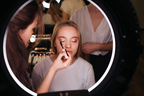 Regardez dans le miroir dans la salle de beauté. Le maquilleur met du mascara sur les cils. Jeune femme blonde assise sur une chaise les yeux fermés. Travailler dur derrière le client . — Photo