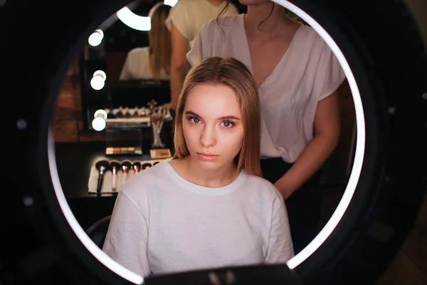 Sérieux jeune modèle blonde regarder dans le miroir dans la salle de beauté. Coiffeur debout derrière et travailler . — Photo