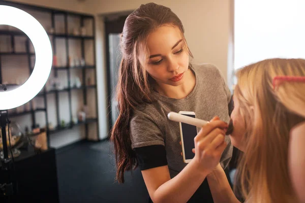 Jeune maquilleuse appliquant le fard à joues sur les joues blondes dans la salle de beauté. Regard professionnel sur le client . — Photo