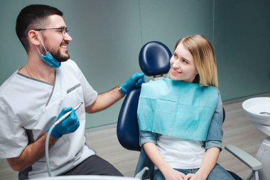 Mutlu genç kadın müşteri diş hekimliği sandalyede oturmak. O bakmak diş hekimi ve gülümseme. Maske ve beyaz genç adam soymak holding makine diş tedavisi için.