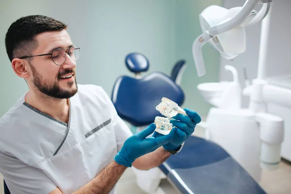 Joven dentista alegre sentarse en la sala de odontología. Sostiene y mira la mandíbula artificial con dientes. Médico usar gafas y sonrisa . — Foto de Stock