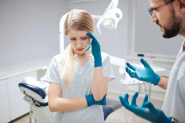 Mujer joven en bata blanca de pie en la habitación con el dentista. Se toma de la mano y sufre de dolor de cabeza. Hombre mira a la mujer y preocupado . — Foto de Stock