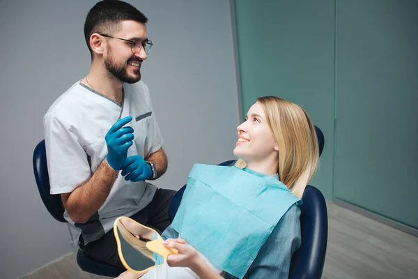 Joyeux dentiste positif et client en dentisterie. Ils se regardent et sourient. Une cliente s'assoit sur une chaise et tient un miroir. Elle a l'air satisfaite . — Photo