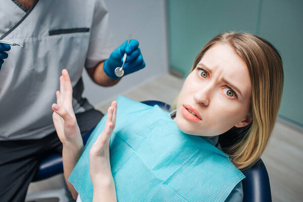 Молодая женщина выглядит напуганной. Она сидит в кресле стоматолога. Женщина смотрит в камеру и отталкивает доктора. Он держит в руках стоматолога
.