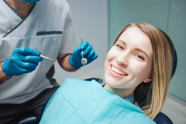 Mujer joven positiva sentarse en la silla de odontología y sonreír a la cámara. Muestra una hermosa sonrisa. Herramientas de sujeción para el tratamiento dental . — Foto de Stock
