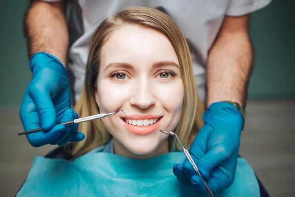 Picutre de buen cliente femenino mirar en la cámara y mostrar los dientes blancos. Hombre médico de pie detrás de ella y mantenga el equipo de dientes cerca de la boca . — Foto de Stock