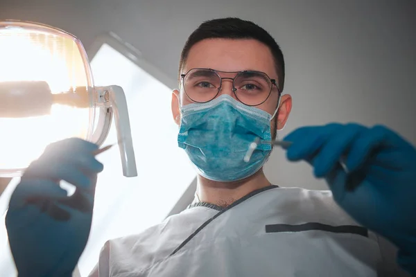 Jovem dentista masculino olhar para a câmera. Ele usa um roupão branco e uma máscara azul. O tipo segura o equipamento dos dentes para tratamento. A lâmpada está ligada. Sala de Odontologia . — Fotografia de Stock