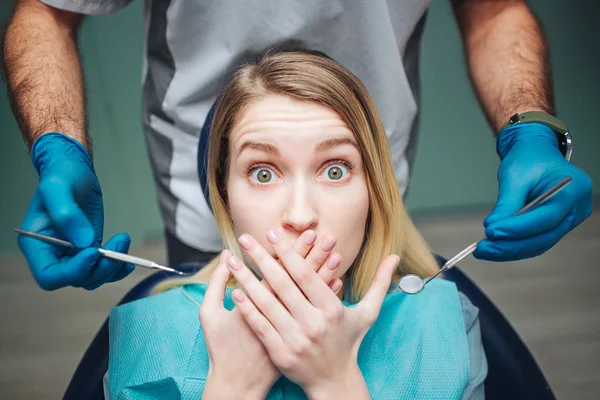 Mujer joven sentada en silla en odontología. Ella tiene miedo y cubre la boca con las manos. El cliente mira a la cámara con miedo en los ojos. Equipo de retención del dentista masculino para el tratamiento de dientes . — Foto de Stock