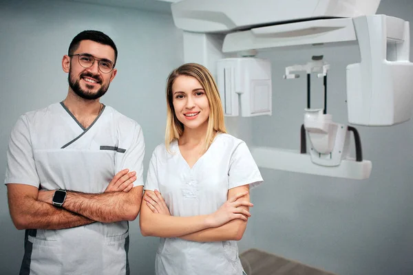 Two dentists stand in x-ray room and pose to camera. They smile and hold hands cossed. Professionals on picture.