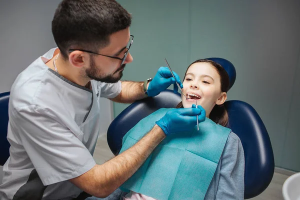 Un dentista joven comprueba los dientes de las chicas. Mantiene la boca abierta. El hombre usa herramientas dentales. Chica sentarse en la silla en la habitación . — Foto de Stock