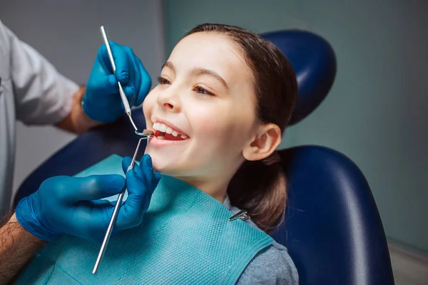 Fille joyeuse assise dans une chaise dentaire dans la chambre. Elle montre des dents. Dentiste faire check-up avec petit miroir et outil . — Photo