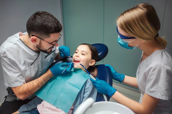 Homme dentiste vérifier les dents des filles avec des outils dentistes. Une assistante se tient à côté. Fille assis dans une chaise dentaire dans la chambre. Elle garde la bouche ouverte . — Photo