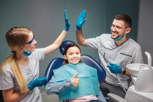 Ung man och kvinna tandläkaren ge high-five till varandra i rummet. De ler. Flickan sitter i stolen och hålla stora tummen upp. Hon ler alltför. — Stockfoto