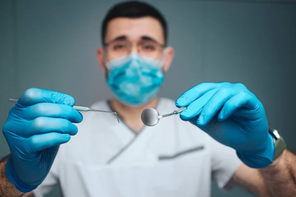 Calma jovens pacíficos fazer dentista em uniforme e máscara. Ele olha para a câmara e segura nas ferramentas dentárias. O tipo usa luvas de látex. Isolado em fundo verde . — Fotografia de Stock