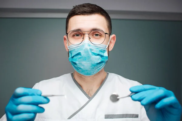 Um dentista jovem e sério usa uniforme e máscara. Ele segura ferramentas dentárias nas mãos com luvas de látex. O tipo olha para a câmara. Isolado em fundo verde . — Fotografia de Stock