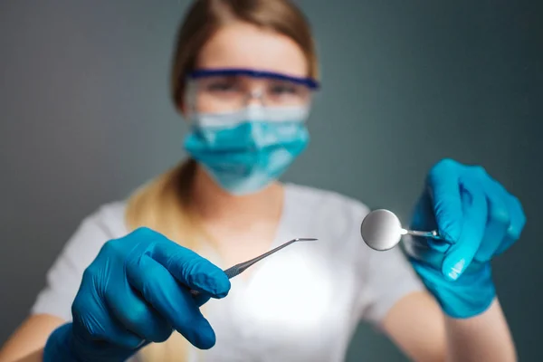 Jovem dentista feminino olhar para a câmera através de óculos. Ela segura as ferramentas dentárias nas mãos. Modelo vestir roupas brancas e máscara azul. Isolado em fundo verde . — Fotografia de Stock