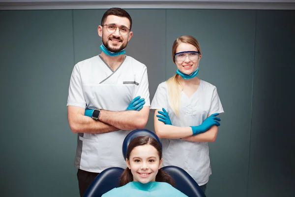 Beau dentiste masculin et féminin positif avec une fille dans une chaise dentaire. Ils ont l'air droit et sourient. Les adultes se tiennent les mains croisées. Isolé sur fond vert . — Photo