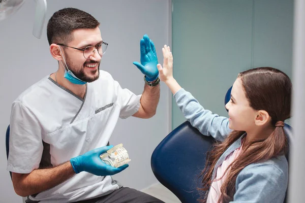 Homme dentiste donnant haute cinq à la fille dans la chaise dentaire. Ils sont dans la chambre. Il a une mâchoire artificielle avec des dents. Heureux et positif . — Photo