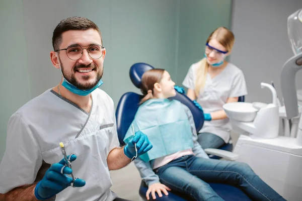 Joyeux dentiste masculin regarder la caméra et sourire. Il tient des outils. Une femme dentiste se tient à la fille. Enfant assis dans une chaise dentaire . — Photo