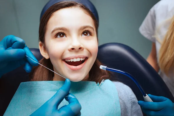 Alegre niño positivo sentarse en la silla dental. Manos en guantes de látex para usar los dientes delanteros. Mujer asisstant stan al lado . — Foto de Stock