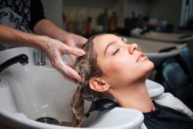 Beautiful young woman sitting near sink while hairdresser washing her hair in beauty salon. Hair stylist at work clipart