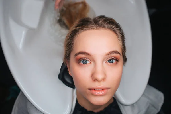 Portrait de belle jeune femme blonde assise sur un lavabo blanc et attendant un coiffeur. Lavage des cheveux dans le salon de beauté — Photo