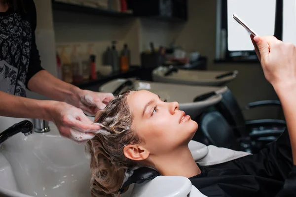 Belle jeune femme regardant téléphone tandis que le coiffeur se lave les cheveux après masque dans un salon de beauté. Concept beauté et personnes — Photo