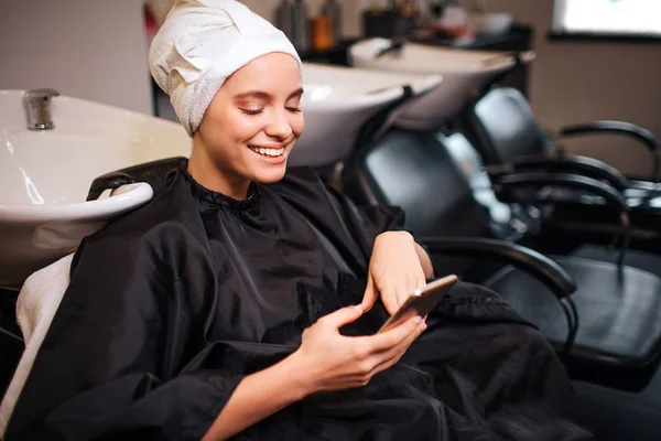 Uśmiechający się piękna kobieta z ręcznikiem na głowie ogląda telefon przed robi fryzurę. Spa włosów w salonie piękności — Zdjęcie stockowe