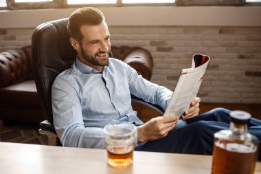 Genç yakışıklı işadamı kendi ofisinde günlüğü okuyun. Tablo ve gülümseme şurada. Pozitif neşeli günlüğü dostum. Cam ve praphene viski masada.