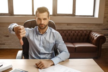Genç ciddi yakışıklı işadamı kendi ofisinde masada otur. O tutmak büyük baş aşağı ve göz ciddi kamerada. Arkasında parlak pencere.