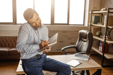 Genç yakışıklı işadamı ve kendi ofisinde telefonda konuşmak masaya oturun. Not defterinde yazmış. İş görüşmesi. Gün ışığından yararlanma penceresinden.