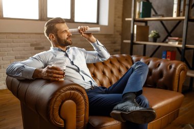 Genç yakışıklı işadamı kanepede oturup kendi ofisinde oynamak. Haddelenmiş günlüğü düz görünüyor. Adam elinde viski ile cam tutun. Başka bir bacak. Güven.