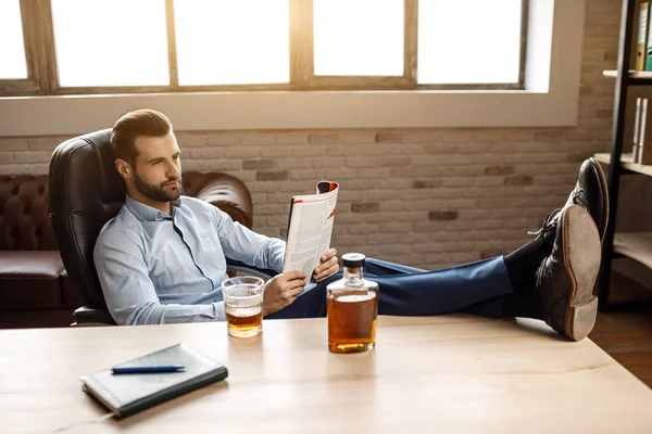 Genç yakışıklı işadamı masaya oturup kendi ofisinde günlüğü okuyun. O masanın üzerinde bacaklarını tut. Cam ve graphene viski ile masada duruyor. Kendinden emin ve güzel.