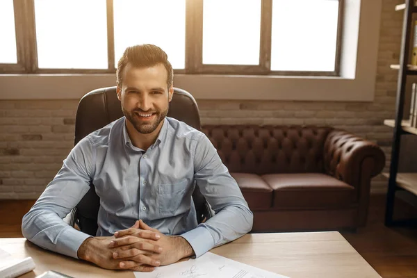 Genç neşeli yakışıklı işadamı masaya oturup kendi ofisinde poz. Doğrudan kameraya bak ve ellerini masanın üstünde tutmak.