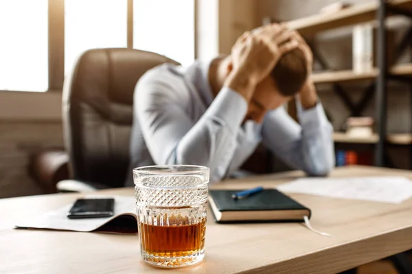 Mladý pohledný podnikatel sedět u stolu a trpí kocovinou ve své vlastní kanceláři. On drží za ruce na hlavu. Sklenice whisky stojí v popředí. — Stock fotografie