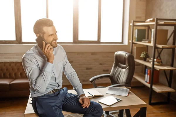 Genç neşeli yakışıklı işadamı ve kendi ofisinde telefonda konuşmak masaya oturun. O gülümseme. İş konuşması. Kendinden emin ve seksi.