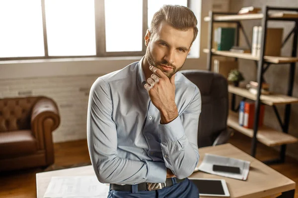 Genç yakışıklı işadamı masada stand ve kendi ofisinde fotoğraf makinesinde oluşturmaktadır. O eli çeneme tut ve düz. Yakışıklı ve kendine güvenen.