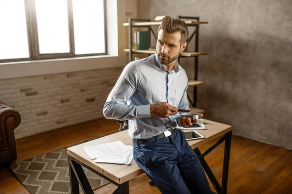 Young handsome businessman hold cigar and pose in his own office. He look to side and sit on table. Confident sexy alfa male. — Stock Photo, Image